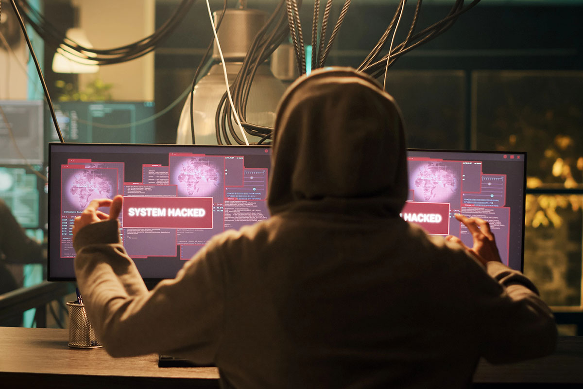 A person in a hoodie viewed from behind, gesturing triumphantly in front of dual computer monitors displaying red "SYSTEM HACKED" warning pop-ups.