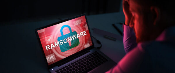 Stressed person sitting in front of a laptop displaying a red "RAMSOMWARE" security warning on the screen.