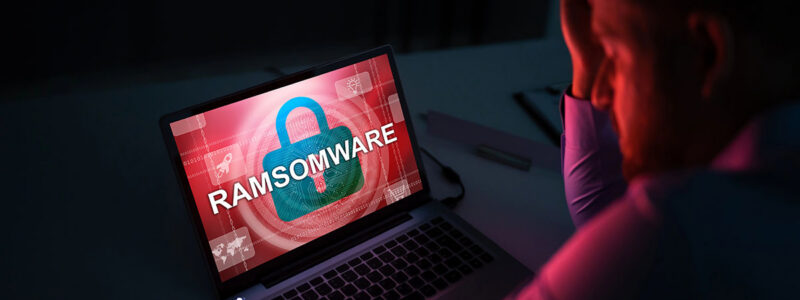 Stressed person sitting in front of a laptop displaying a red "RAMSOMWARE" security warning on the screen.