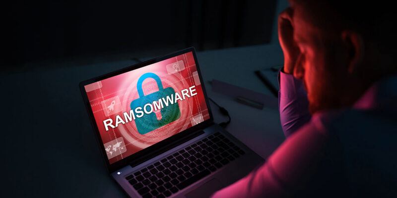 Stressed person sitting in front of a laptop displaying a red "RAMSOMWARE" security warning on the screen.