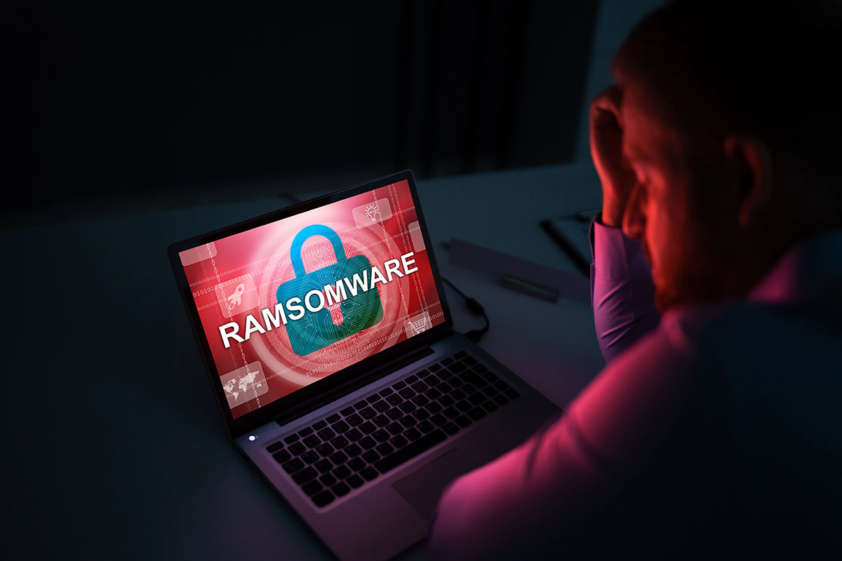 Stressed person sitting in front of a laptop displaying a red "RAMSOMWARE" security warning on the screen.